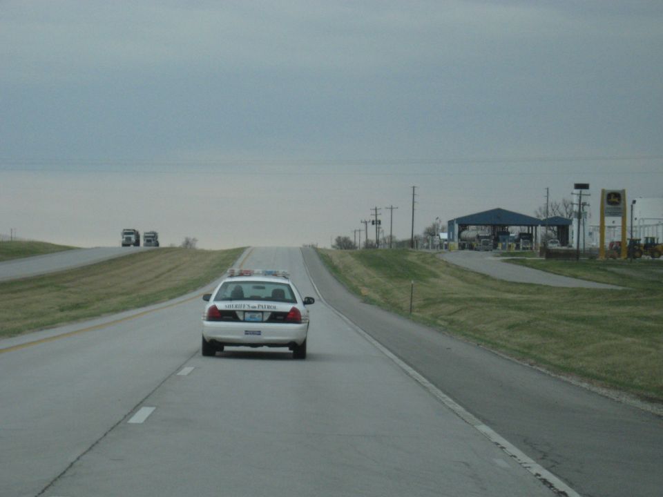 Police car driving on a highway, with a building and some equipment in the background