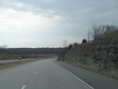1978 Ford Bronco - Two cars travel down a highway under a cloudy sky, with a wooded area and a rocky hillside visible.
