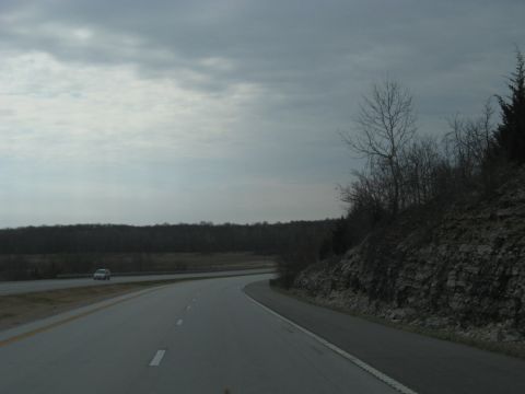 1978 Ford Bronco - A lone car travels down a highway bordered by trees and a rocky embankment under an overcast sky.