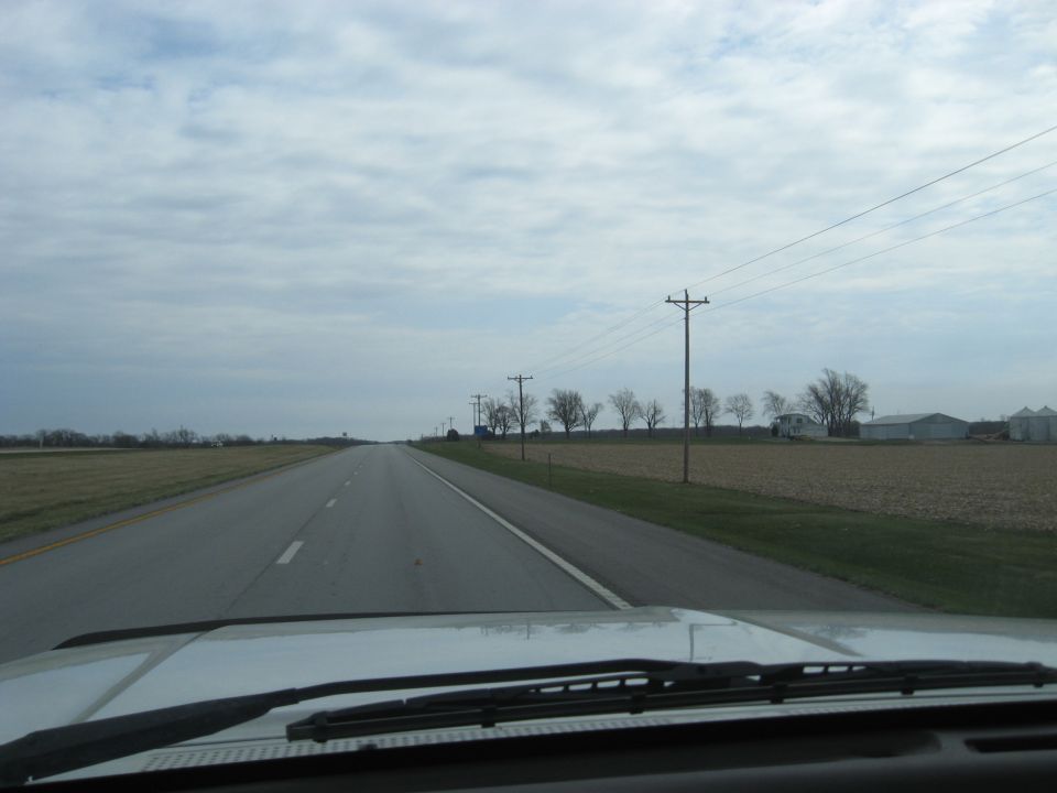 View from inside a vehicle looking out at a highway stretching into the distance, with fields and telephone poles lining the road