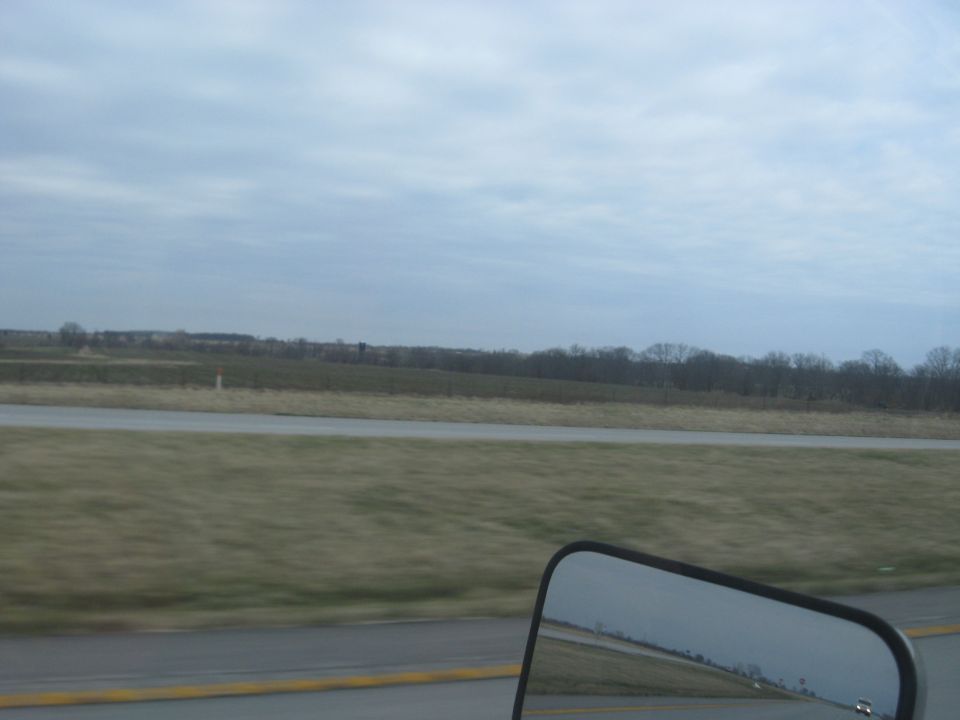 view of a highway with fields and trees under a cloudy sky, seen from a vehicle's side mirror