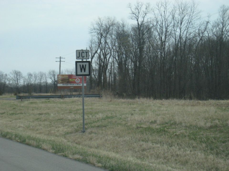 JCT W road sign with a billboard advertising fast food in the background, surrounded by bare trees