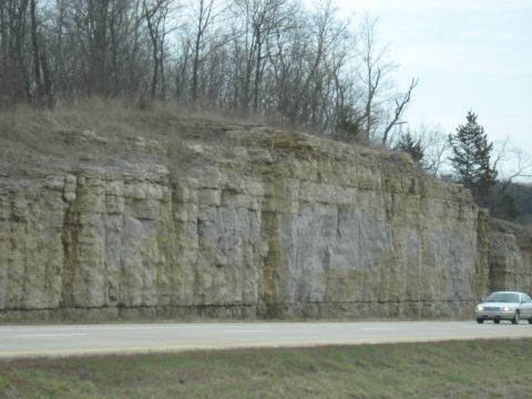 1978 Ford Bronco - A silver sedan travels along a highway adjacent to a towering rock formation under a cloudy sky.