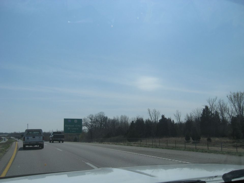 Two trucks on a highway under a blue sky. A sign indicates Kansas City and St. Louis