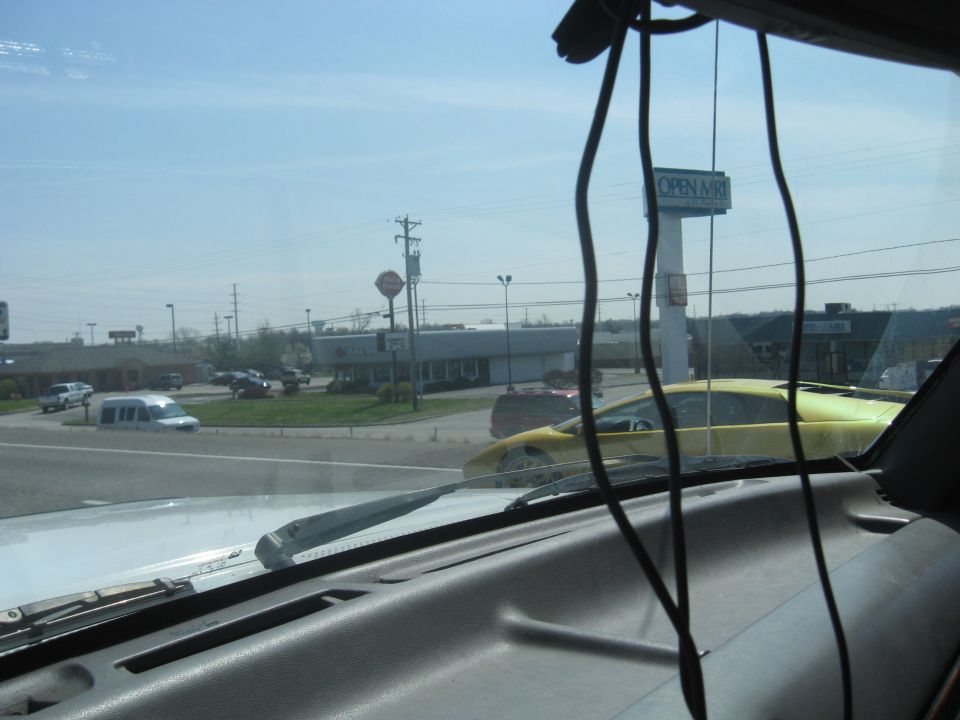 Yellow sports car seen from inside a vehicle, with other cars and buildings in the background