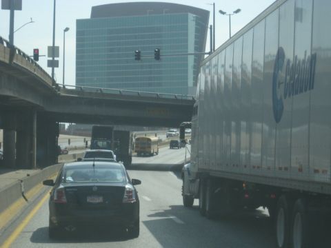 1978 Ford Bronco - A black sedan is seen from behind on a highway, with a large semi-truck to its right and a modern building in the background.