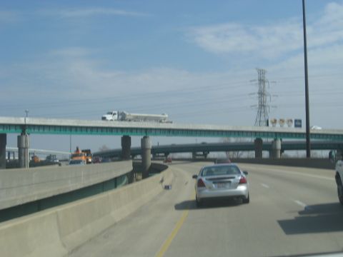 1978 Ford Bronco - A silver sedan drives on a highway under an overpass with a truck on it.