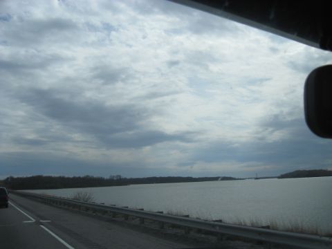 1978 Ford Bronco - A dark SUV travels down a highway beside a wide expanse of water on an overcast day.