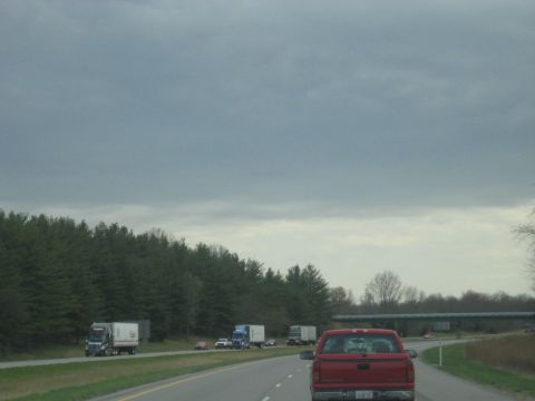1978 Ford Bronco - A red pickup truck travels down a highway, passing other vehicles and a line of trees under a cloudy sky.