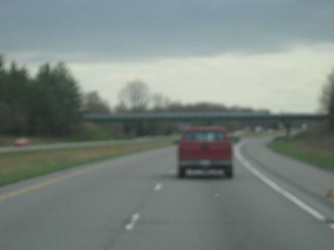 1978 Ford Bronco - A red pickup truck travels down a highway, with trees and an overpass visible in the background.