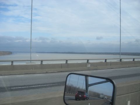 1978 Ford Bronco - A red SUV is seen in the side mirror of a vehicle traveling on a bridge, with a large river or lake stretching to the horizon.