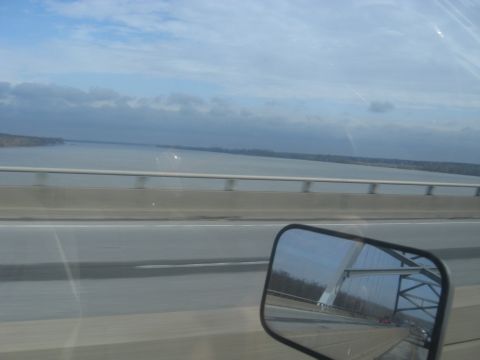 1978 Ford Bronco - Driving across a bridge over a large body of water, with a reflection of the bridge structure in the side mirror.
