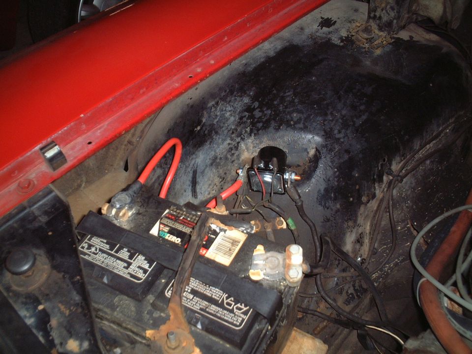 Battery and solenoid in a red vehicle's engine bay with red positive cable and corroded terminals.