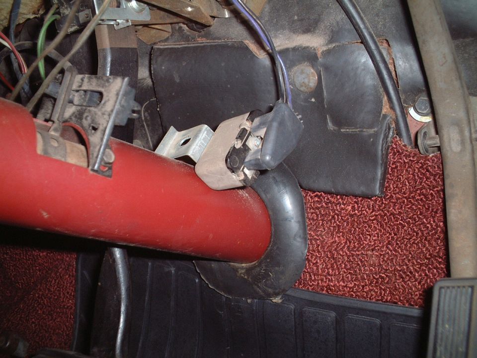 Underdash view of red steering column with switch and wiring above dark red carpet and floor mat