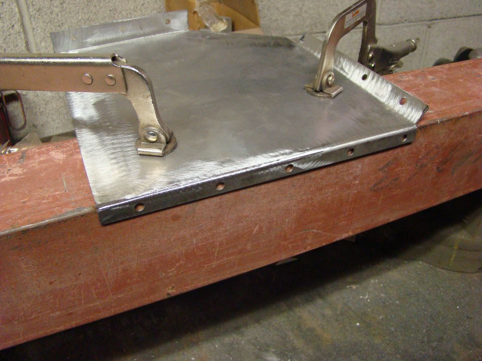 Sheet metal piece with drilled holes, held by clamps to a red wooden beam