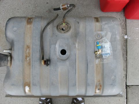 1979 Ford Bronco - A metal fuel tank, likely for a vehicle, is shown with attached hoses and a fuel filter, resting on a concrete surface.