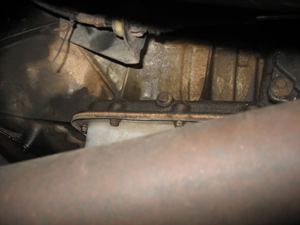 Transmission pan with bolts and casing, showing oil stains and a rusty pipe in the foreground