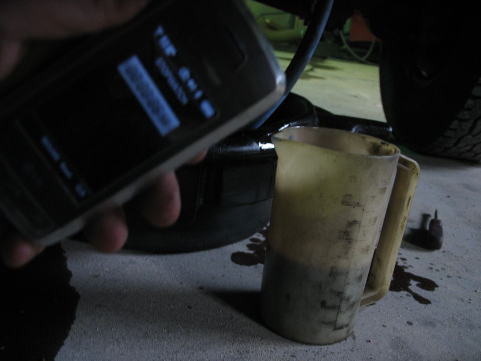 Phone screen shows "THE QUIK STOPWATCH" and a barcode. A measuring jug with dark fluid sits on a concrete floor next to spilled oil