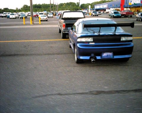 1990 Ford Taurus - A blue sports coupe with a large rear spoiler is parked in a lot, with a pickup truck beside it.