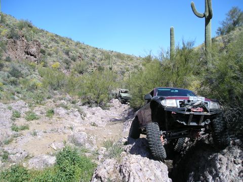 1986 Toyota Hilux - Two modified trucks navigate a challenging rocky path in a desert landscape under a clear blue sky.
