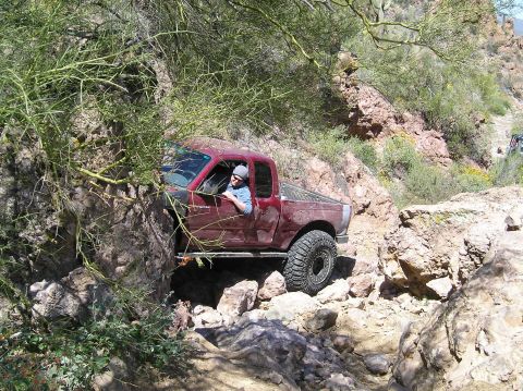 1986 Toyota Hilux - A maroon pickup truck maneuvers through a tight, rocky off-road path, with another vehicle visible in the distance.