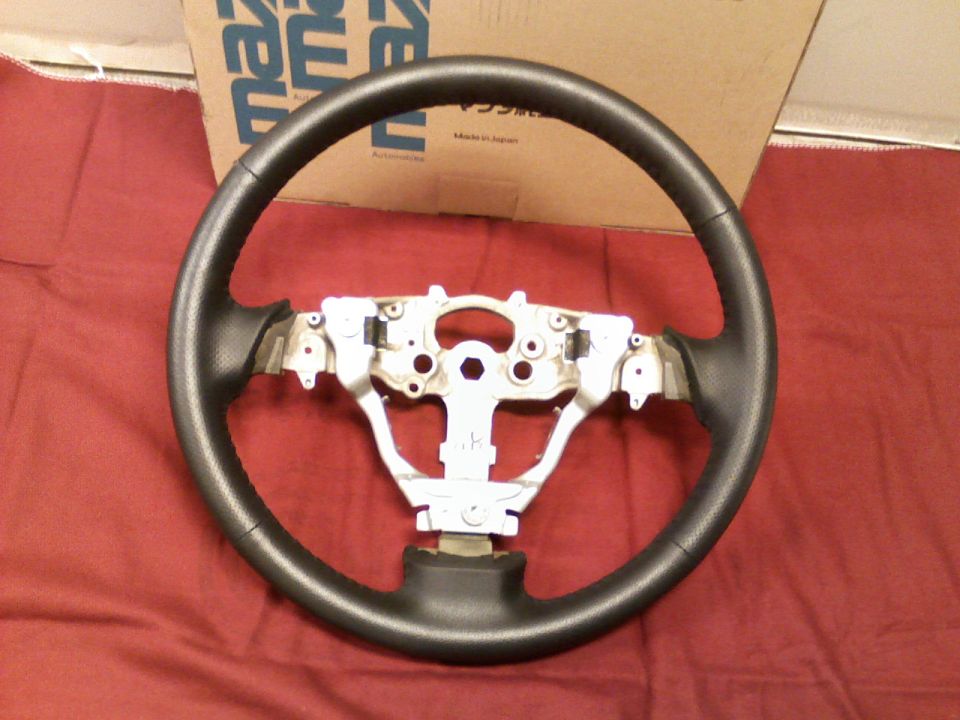 Black steering wheel without airbag, exposed silver bracket, on red cloth near a Mazda box.