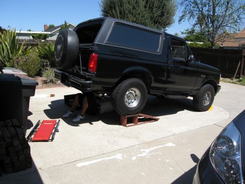 1986 Ford Bronco - A person works on the rear shocks of a 1986 Ford Bronco lifted on ramps in a driveway.