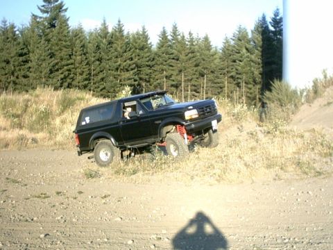 1995 Ford Bronco - A black SUV navigates an off-road trail, its lifted suspension against a backdrop of pine trees.