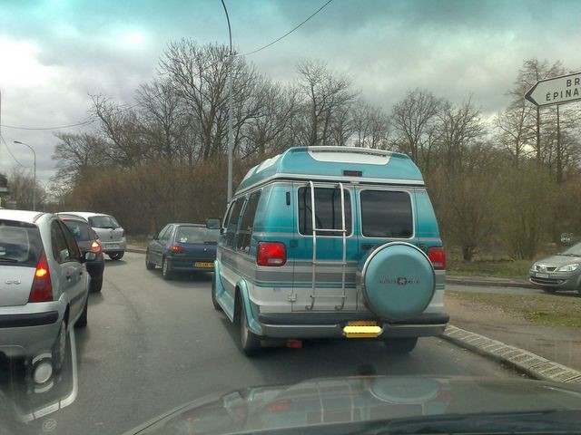 Blue and silver van with a ladder on the back and a spare tire cover, seen from behind in traffic