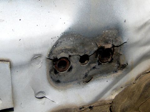 1996 Ford Bronco - Close-up of a white sheet-metal panel with a rusted, burned-through mounting boss and two adjacent holes, the paint scorched black around a…