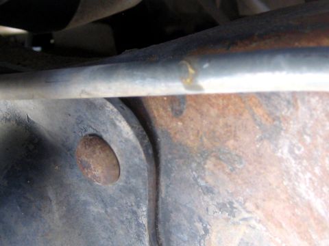 1996 Ford Bronco - Extreme close-up underneath a truck showing heavily rust-scaled frame or crossmember metal, with a plug or rivet head in a stamped bracket…