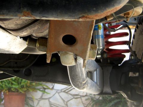 1996 Ford Bronco - Underbody shot of a surface-rusted fabricated bracket bolted to the frame with large yellow-zinc hardware, a bright bare-metal exhaust pipe…