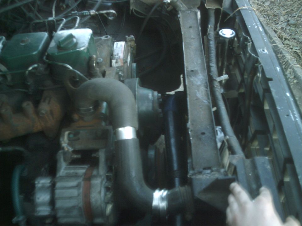Engine bay with hoses, radiator, and alternator visible. A hand is near the radiator