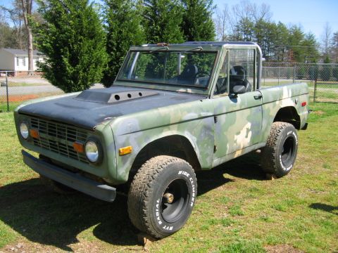 1992 Ford Mustang - Three-quarter front view of an early Ford Bronco in patchy green camouflage paint, parked on grass with the top and doors removed and a rol…