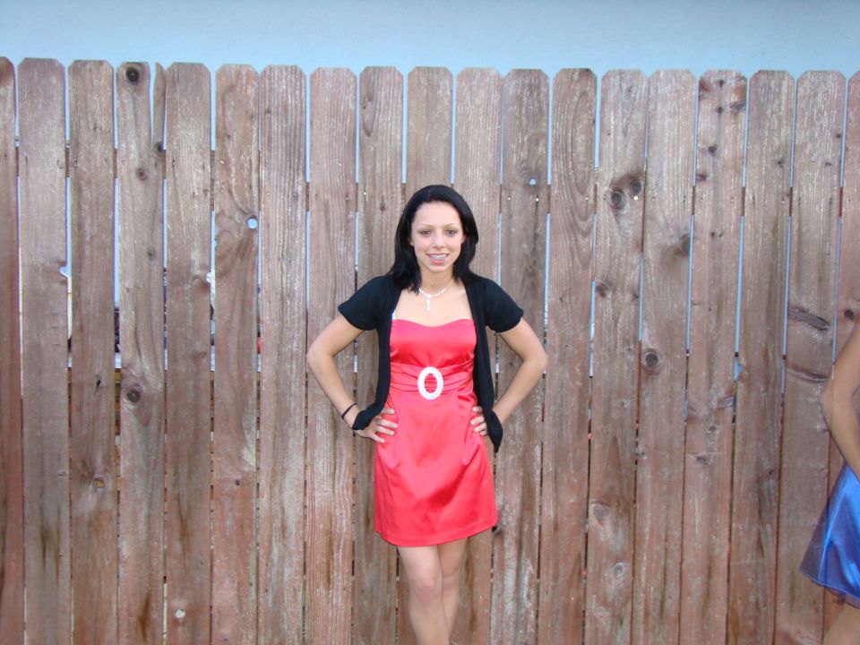 Woman in red dress and black shrug standing in front of a wooden fence