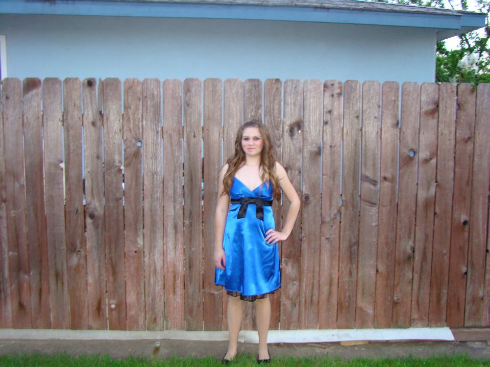Young woman in a blue satin dress with a black bow stands in front of a wooden fence