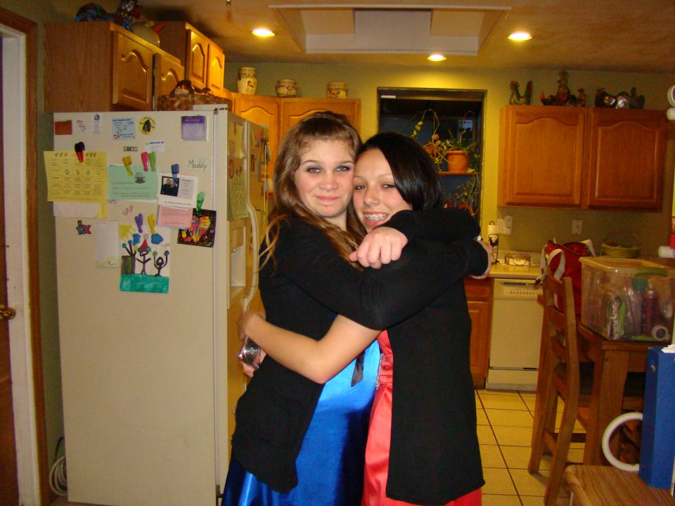 Two young women in formal dresses hug in a kitchen with a refrigerator covered in notes