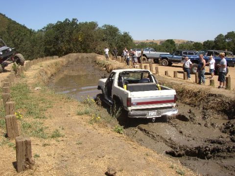 1989 Ford Bronco - A white SUV navigates a muddy obstacle course with spectators watching in the background.