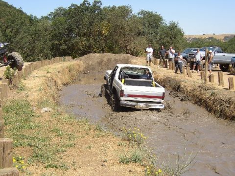 1989 Ford Bronco - Off-road vehicles navigate a muddy track with onlookers watching from the side.