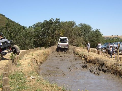 1989 Ford Bronco - Off-road vehicles navigate a muddy track under a clear blue sky, with spectators and parked trucks nearby.