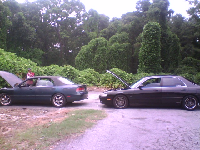 Two sedans, one green and one black, parked facing each other with hoods open near kudzu-covered woods