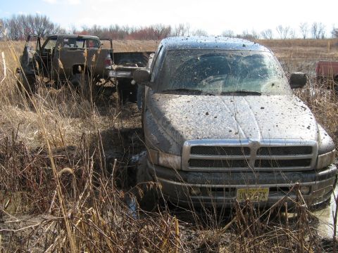 1990 Ford Bronco - Two pickup trucks are stuck in the mud in a field with dry grass and sparse trees.