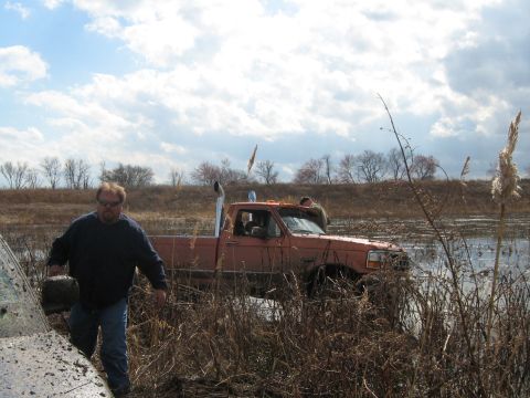 1990 Ford Bronco - A man walks past a stuck orange pickup truck in a muddy, watery field under a cloudy sky.