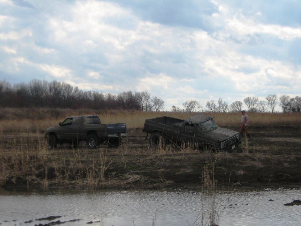 Two trucks stuck in mud, one being pulled by the other. A person stands nearby