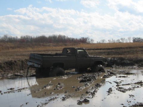 1990 Ford Bronco - A dark-colored pickup truck is deeply stuck in thick mud and water under a cloudy sky.
