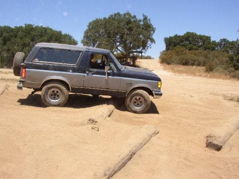 1989 Ford Bronco - A two-tone Ford Bronco navigates an off-road course with logs placed on the sandy terrain.