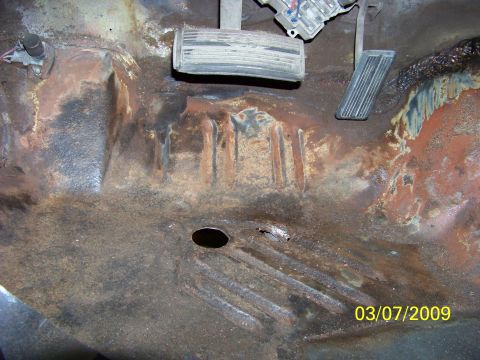 1974 Dodge Challenger - The interior floor pan of a car shows extensive rust and damage, with visible pedals and a hole in the metal.