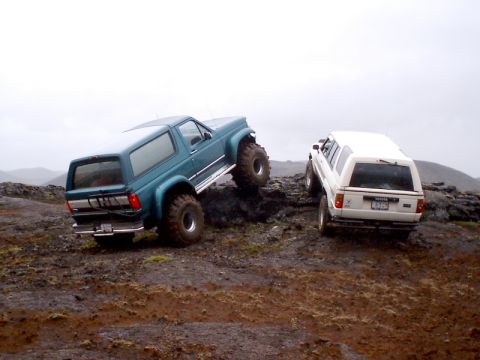 1996 Ford Bronco - Two off-road vehicles, a teal Ford Bronco and a white Toyota SUV, navigate a rocky, muddy landscape under an overcast sky.