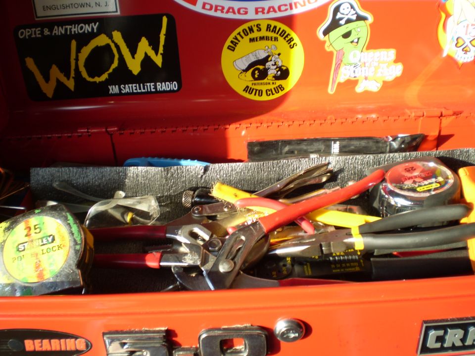 Tool box drawer of pliers and tape measures under a lid covered in racing and band stickers