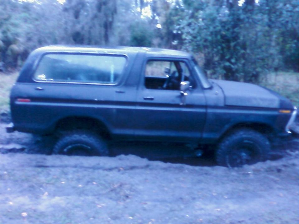 Dark SUV stuck in mud, side view. Visible front fender, door, rear quarter panel, and tire. Mud covers the lower body